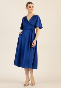 Kuningan biru royal yang dibungkus dengan lengan pendek berbentuk sayap, pinggang yang terikat, saku samping, dan rok mengalir yang jatuh di bawah lutut.