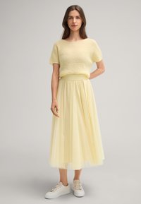 Gelbes gestricktes Kurzarmoberteil kombiniert mit einem fließenden gelben Tüll-Midirock und weißen Leder-Sneakern. Texturiertes Outfit in sanften Farben.