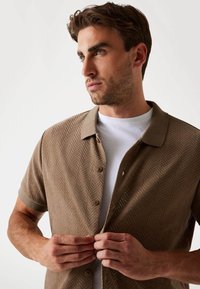 Chemise tricotée marron à manches courtes avec un motif en chevrons, fermeture à boutons et un col contrasté sur un T-shirt blanc.