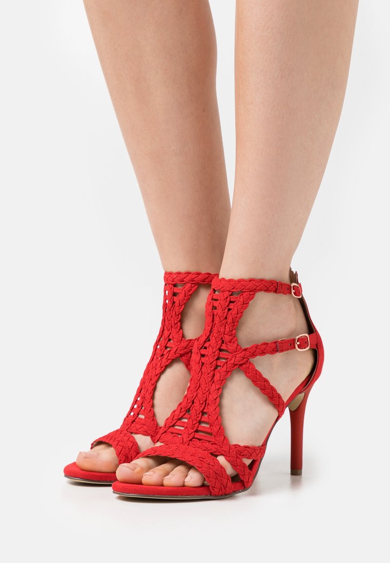 Pieds chaussés de sandales à talons rouges avec des brides tressées et des doubles boucles à la cheville, présentés sur un fond blanc uni.