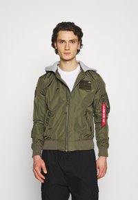 Olivgrüne Bomberjacke mit grauer Kapuze, Reißverschluss, gerippten Bündchen, Seitentaschen und einem roten Etikett Detail am Ärmel. Strukturiert.