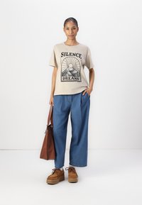 Beige t-skjorte med "SILENCE DREAMS" grafikk, kombinert med løse blå denimbukser og brune platåsko. Brun veske slengt over skulderen.