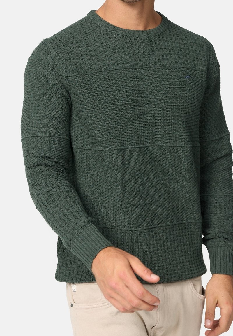 Homme portant un pull en tricot texturé vert foncé et un pantalon beige clair, la main gauche près de la taille et la main droite tendue vers l'avant.