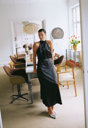 Une femme porte une robe noire à pois et à dos nu, nouée autour du cou, dans une salle à manger lumineuse avec des chaises en bois, une table et des fleurs.