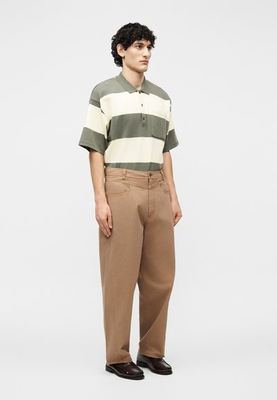 Άνδρας με σγουρά μαλλιά, φορώντας υπερμεγέθη μπεζ παντελόνια, πράσινο-λευκή κοντομάνικη πόλο μπλούζα με ρίγες και σκούρα καφέ παπούτσια, στέκεται πλάγια.