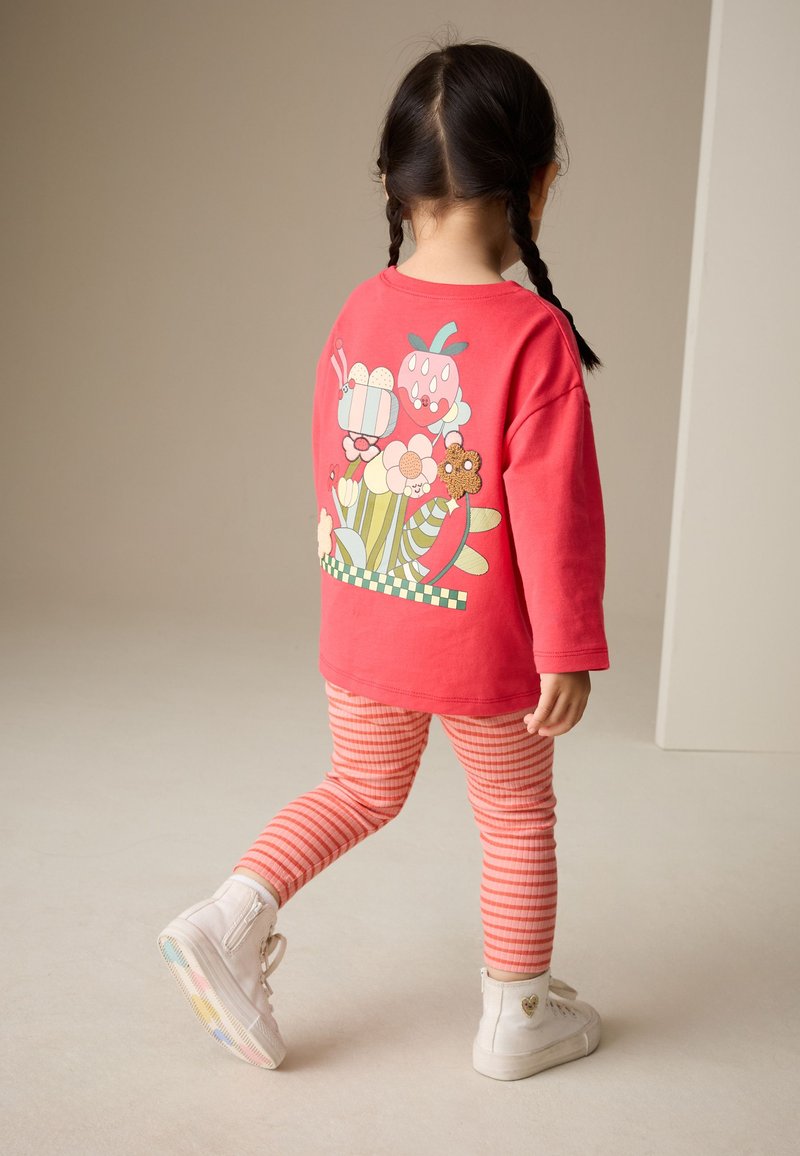 Dijete s pletenicama u crvenoj majici dugih rukava s šarenim cvjetnim i insektnim uzorcima, prugastim tajicama i bijelim tenisicama koje hodaju u zatvorenom prostoru.