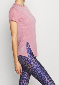 Woman wearing a pink short-sleeve mesh top and blue and purple leopard print leggings, standing sideways against a plain background.
