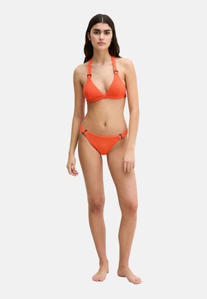 Kobieta stojąca na boso, ubrana w pomarańczowe bikini na szyję z teksturą oraz metalowymi pierścieniami na ramiączkach i bokach, neutralne tło.