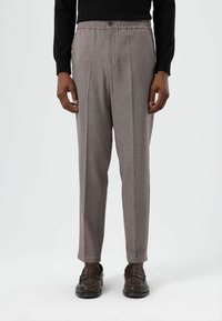 Grijze broek met een textuur, een ontspannen pasvorm en plooien, voorzien van een knoopsluiting en een elastische tailleband. Draagt donkerbruine loafers.