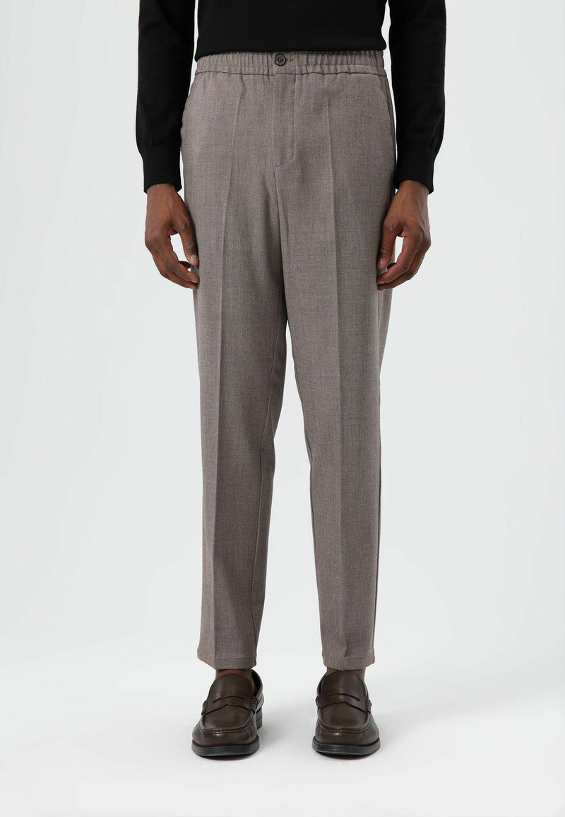 Grijze broek met een textuur, een ontspannen pasvorm en plooien, voorzien van een knoopsluiting en een elastische tailleband. Draagt donkerbruine loafers.