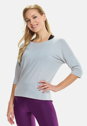 Helles graues Oberteil mit Dreiviertelärmeln aus weichem Stoff; verfügt über einen runden Ausschnitt und ein dezentes Logo-Detail. Kombiniert mit lila Leggings.