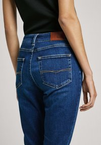 Modré rifle z denimu s priliehavým strihom, s hnedou koženou nášivkou a kontrastným žltým prešívaním na zadných vreckách.