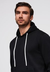 Černá mikina s kapucí, která má přední klokánkovou kapsu, bílé šňůrky a texturovanou tkaninu. Jednoduchý design s volným střihem.