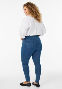 Jeans de mezclilla azul de cintura alta con ajuste skinny, que cuentan con dos bolsillos traseros y una textura suave, combinados con una blusa blanca suelta.