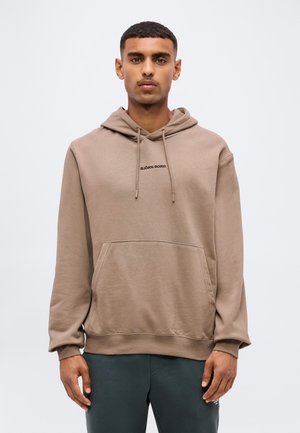 Jeune homme portant un sweat à capuche Björn Borg marron clair avec une poche avant et un pantalon foncé, debout devant un fond clair uni.
