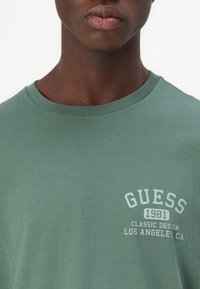 Zöld pamut póló, amelynek kerek nyakrésze és fehér grafikai nyomata van, amelyen a "GUESS 1981 CLASSIC DESIGN LOS ANGELES, CA." felirat látható.