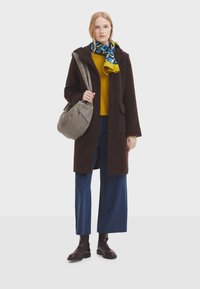 Cappotto di lana marrone, maglione di cotone giallo, pantaloni a gamba larga blu, sciarpa con motivo blu/verdi/giallo, borsa grigia con dettaglio della cerniera, stivali marroni.