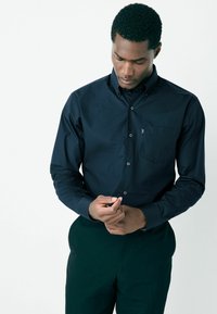 Sötétkék gombos ing pamut anyagból, hosszú ujjal, szabványos gallérral és mellzsebbel. A modell a sötét nadrágot viselve igazítja az ing mandzsettáját.