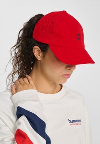 Rød cap med perforeret design; buet skygge og sort logoaccent. Bæres med en hvid skjorte med blå og røde stribedeærmer.