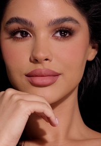 Primer plano de un rostro con un tono de piel suave y medio, labios mate malva llenos, cejas definidas y cabello oscuro, mano descansando en el mentón.