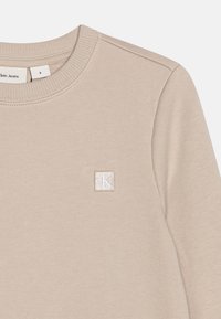 Felpa beige realizzata in tessuto morbido con scollo rotondo e polsini a costine. Presenta un patch a forma di quadrato con il logo CK sul lato sinistro del petto.
