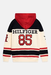 Huppari, jossa on kermainen pohjaväri, laivastonsiniset ja punaiset raidat, suuret "HILFIGER"-kirjaimet ja punaisella oleva "85". Pehmeä materiaali, normaali istuvuus.