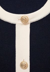 Cardigan en maille marine avec une patte de boutonnage crème et des boutons dorés embossés, mettant en valeur une couture texturée et des couleurs contrastées.