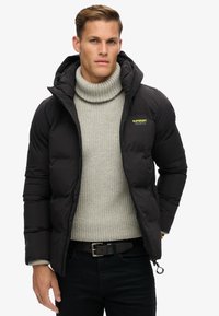 Schwarze Puffjacke aus glänzendem Stoff mit Kapuze. Darunter ist ein strukturierter grauer Pullover zu sehen, sowie ein sichtbares Logo in Neon-Gelb.