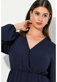 Marineblaue Kleid mit tiefem V-Ausschnitt, elastischem Bund und voluminösen langen Ärmeln. Glatter Stoff mit zarter Textur. Sichtbare silberne Ohrringe.