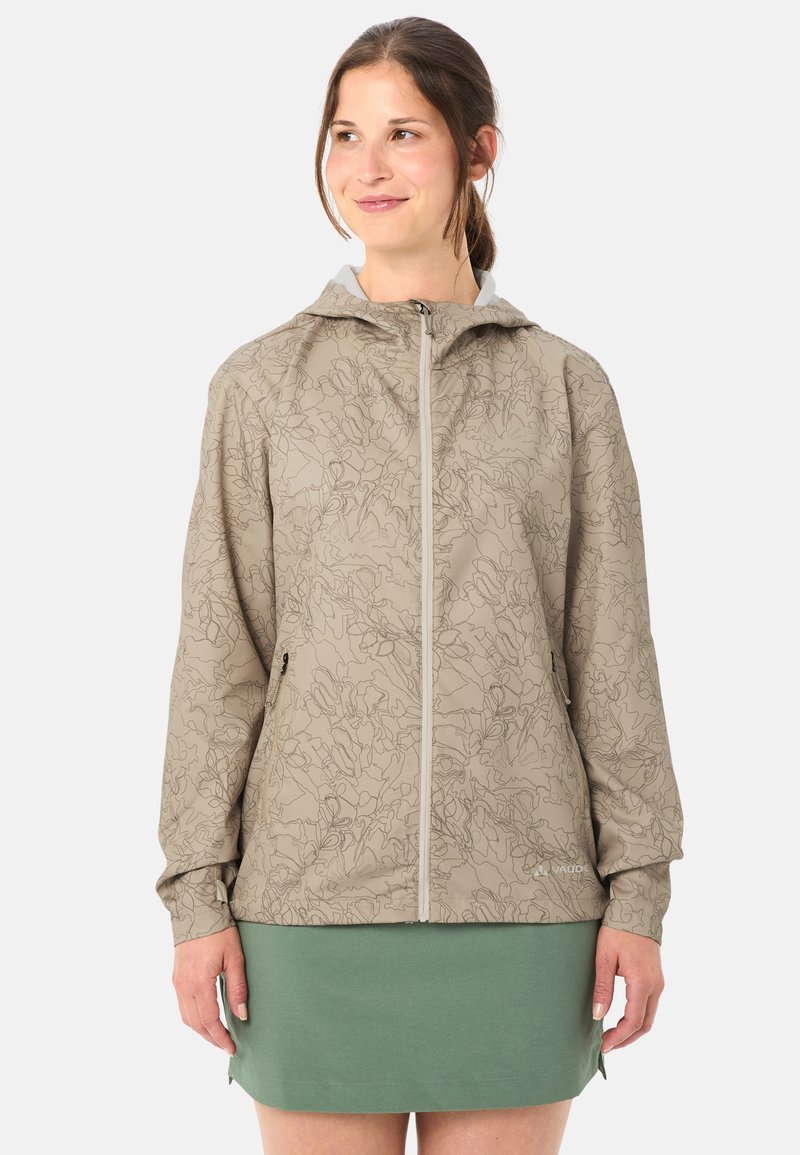 Giovane donna in piedi che indossa una giacca beige con cappuccio e zip, decorata con un motivo astratto, e una gonna verde su sfondo bianco.