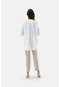 Weiße bestickte Bluse mit langen, puffigen Ärmeln und plissiertem Rücken, kombiniert mit beigen Hosen und hellen Schuhen.