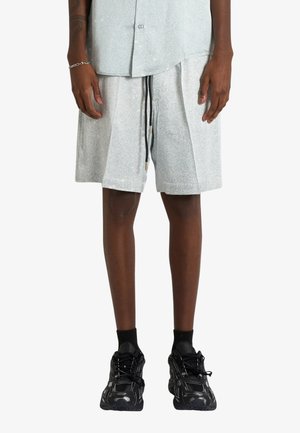Persona che indossa pantaloncini grigio chiaro con coulisse nera, una maglietta abbinata, calzini neri e scarpe da ginnastica nere, in piedi su uno sfondo semplice.