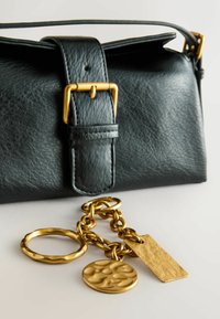 Must must läbipaistev nahk kott, millel on struktureeritud disain; sellel on kullast pandla ja ahel, kus on kolm dekoratiivset talismanit, sealhulgas tekstureeritud ring ja ristkülikukujuline silt.