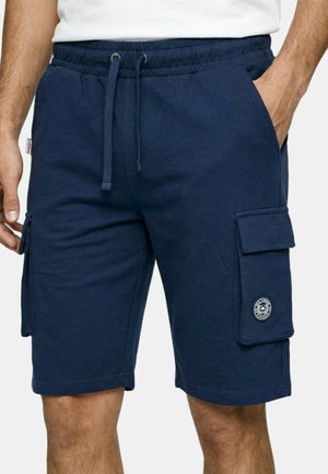 Homme portant un short cargo bleu marine avec taille à cordon et poches à rabat, debout avec une main dans la poche.