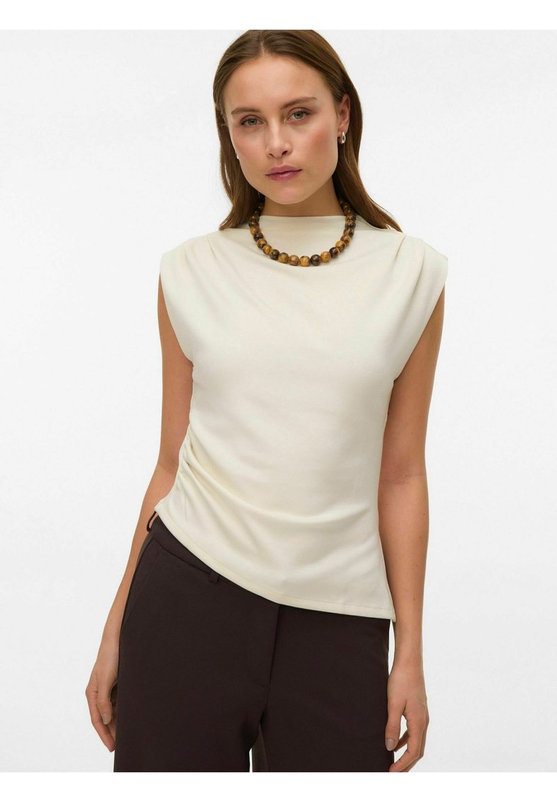 Top senza maniche color crema con collo alto, realizzato in tessuto morbido e liscio con dettaglio arricciato sul lato, abbinato a pantaloni scuri.