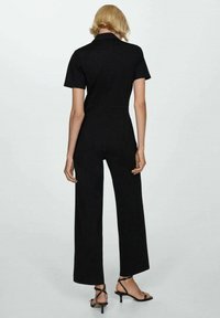Sort jumpsuit med en struktureret design, kortærmede, og brede ben. Har en krave og en glat tekstur. Bæret med sorte strophæle.