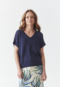 Maglione lavorato a maglia navy con scollatura a V, maniche corte e orlo a coste; abbinato a una gonna color crema con motivi a foglia verdi e blu.
