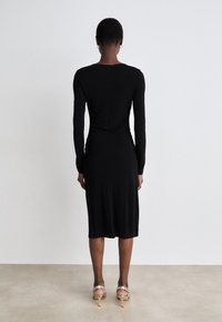 Black long-sleeve dress with fitted silhouette and back zip. Smooth texture, knee-length, worn with strappy metallic-heeled sandals.