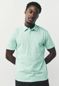 Světlý zelený polo tričko s krátkým rukávem, které má geometrický vzor, zapínací límeček a jemnou strukturu látky. Nosené neformálně s černými kalhotami.