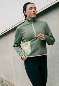 Casaco com fecho éclair verde claro, com padrão texturizado, colarinho alto e um bolso verde claro em contraste. Combinado com leggings pretas ajustadas.