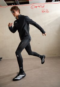 Giovane uomo con abbigliamento sportivo nero Adidas e occhiali da sole che corre al chiuso contro una parete liscia su un pavimento liscio.