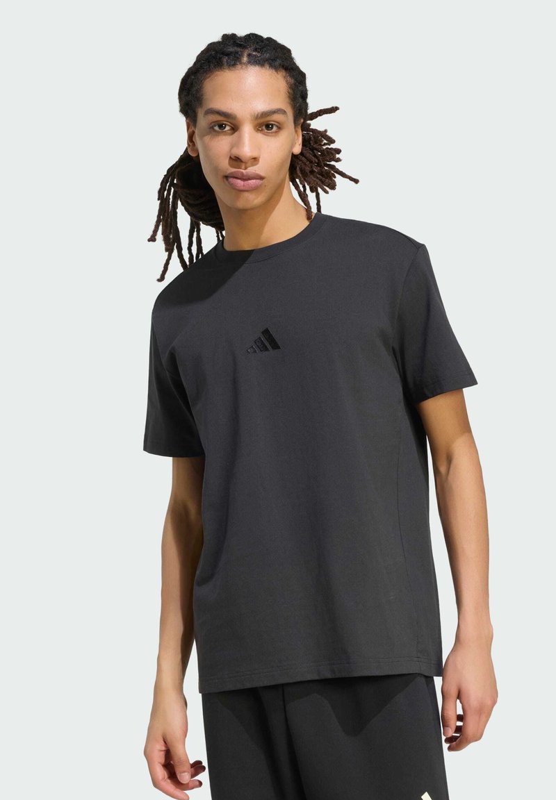 Jeune homme avec des dreadlocks portant un t-shirt Adidas noir et un pantalon noir, debout devant un fond gris clair.