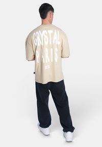 Béžové oversized tričko s výrazným bílým textem "CRYSTAL PARIS" na zádech, spárované s černými kalhotami a bílými teniskami.