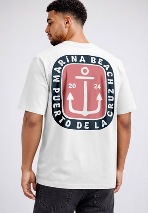 Man wearing white T-shirt with a large pink and navy anchor design and text "Marina Beach Puerto de la Cruz 2024" on the back.