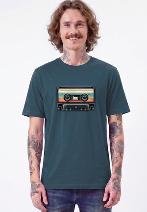 Jonge man met krullend haar en tatoeages die een donker turquoise T-shirt draagt met een kleurrijk vintage cassettebandje ontwerp, glimlachend tegen een witte achtergrond.