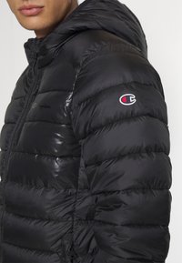 Gros plan d'une personne portant une doudoune noire Champion avec capuche, logo visible sur la manche et fermeture éclair à l'avant.