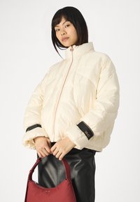 Giacca di piuma crema con colletto alto, zip frontale, dettagli neri sui polsini, indossata con una gonna di pelle nera, mentre tiene una borsa rossa.