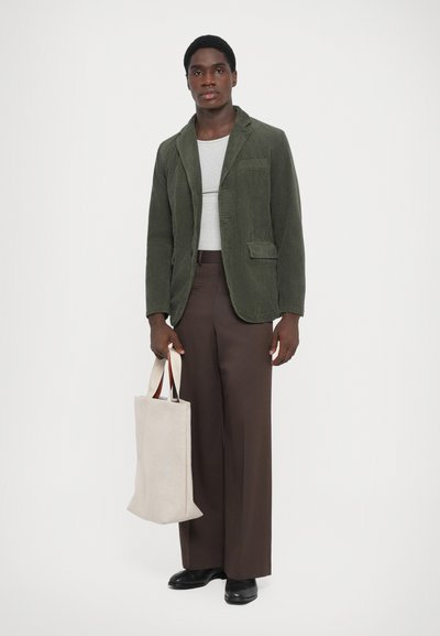 Blazer din catifea în verde măslinie, c�ămașă gri, pantaloni larghi maro, purtând o geantă din material gri cu mânere în contrast, pantofi negri.