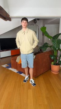 Νεαρός άνδρας στέκεται σε εσωτερικό χώρο φορώντας μπεζ μπουφάν, τζιν σορτς και αθλητικά παπούτσια, με καφέ καναπέ, μεγάλο πράσινο φυτό και τηλεόραση στο φόντο.