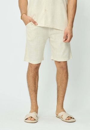 Homme portant un short en lin beige, une chemise assortie, des sandales à glissière beige et un bracelet en or, se tenant avec une main dans la poche.